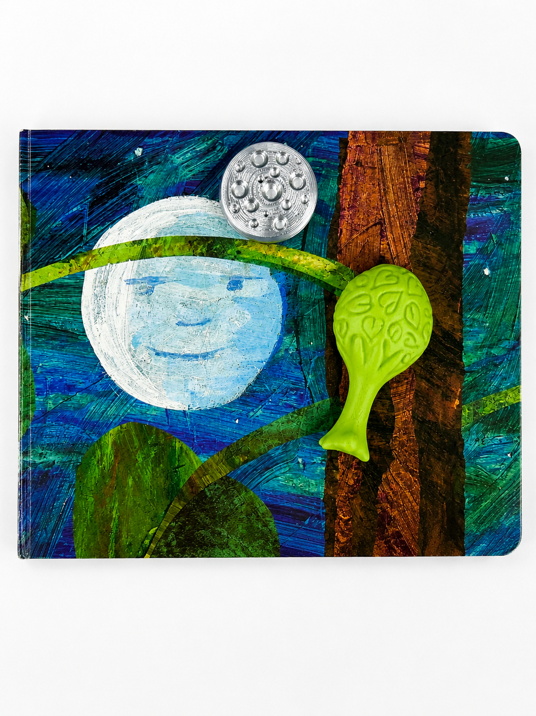 A square decorative item featuring a colorful artistic design with a blue and green textured background, a white moon face, a brown tree trunk, a green leaf-like shape, and a silver circular emblem near the top.