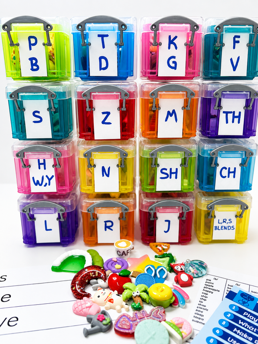 A colorful alphabet sound sorting toy set with 16 clear plastic storage boxes labeled with letters and a pile of assorted plastic sound-effect and rhyming game pieces in front, photographed on a white background.