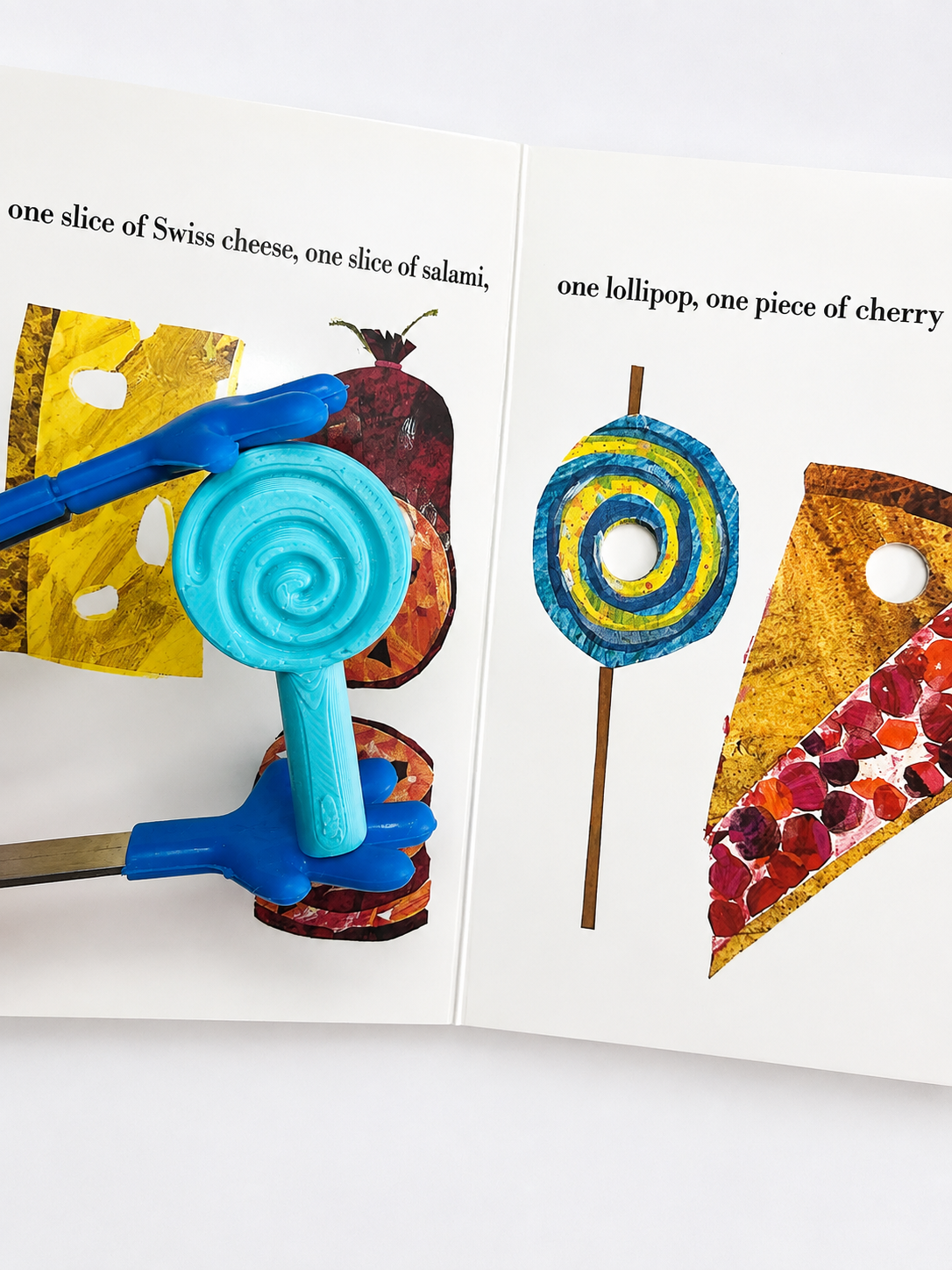 A children's activity book or craft book spread open on a white background, showing printed illustrations of food and blue plastic tools held in hands. The visible text includes 'one slice of Swiss cheese, one slice of salami,' and 'one lollipop, one piece of cherry.