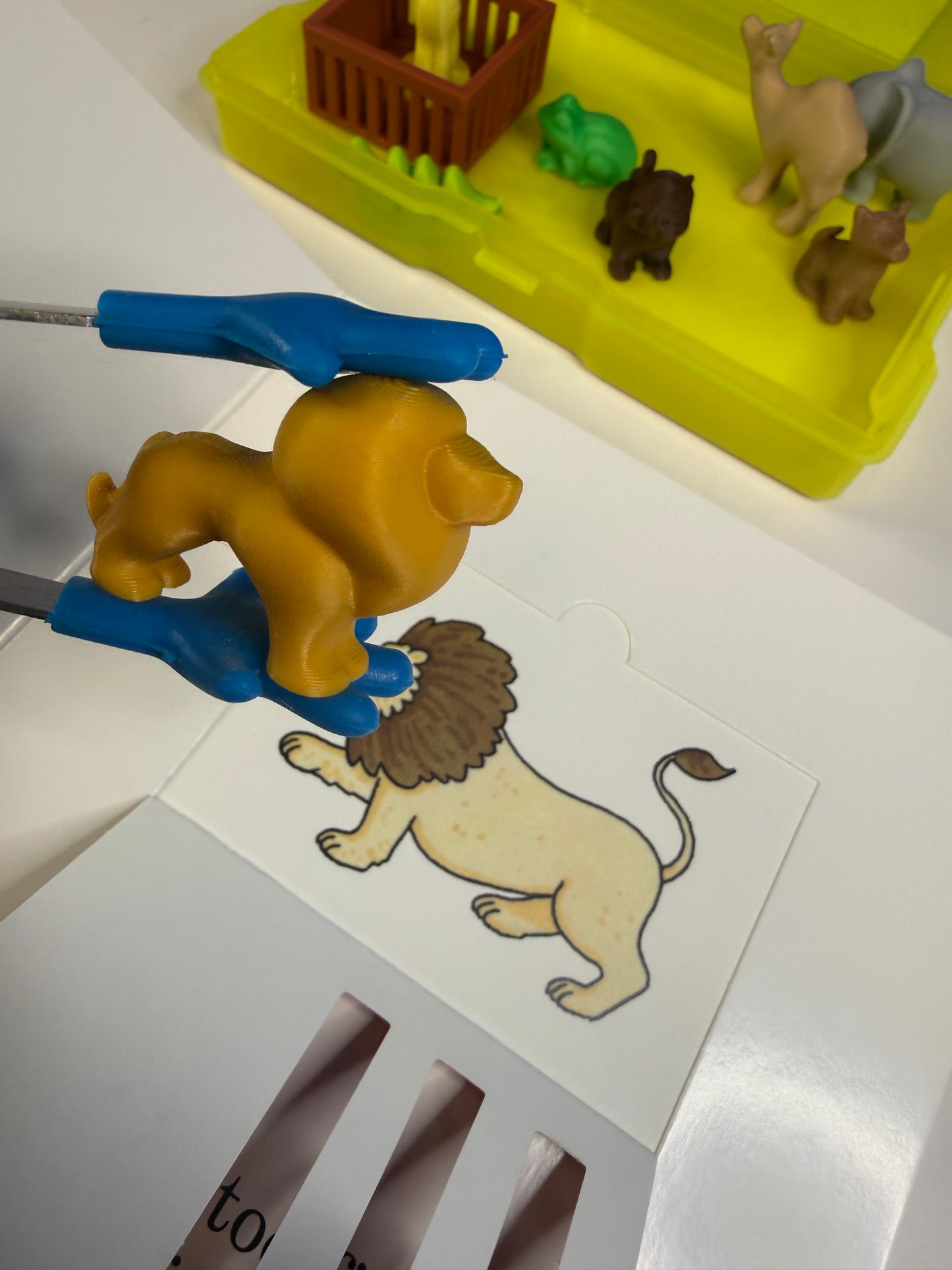 Toy lion figure held by blue plastic clips next to a lion illustration on a white surface.