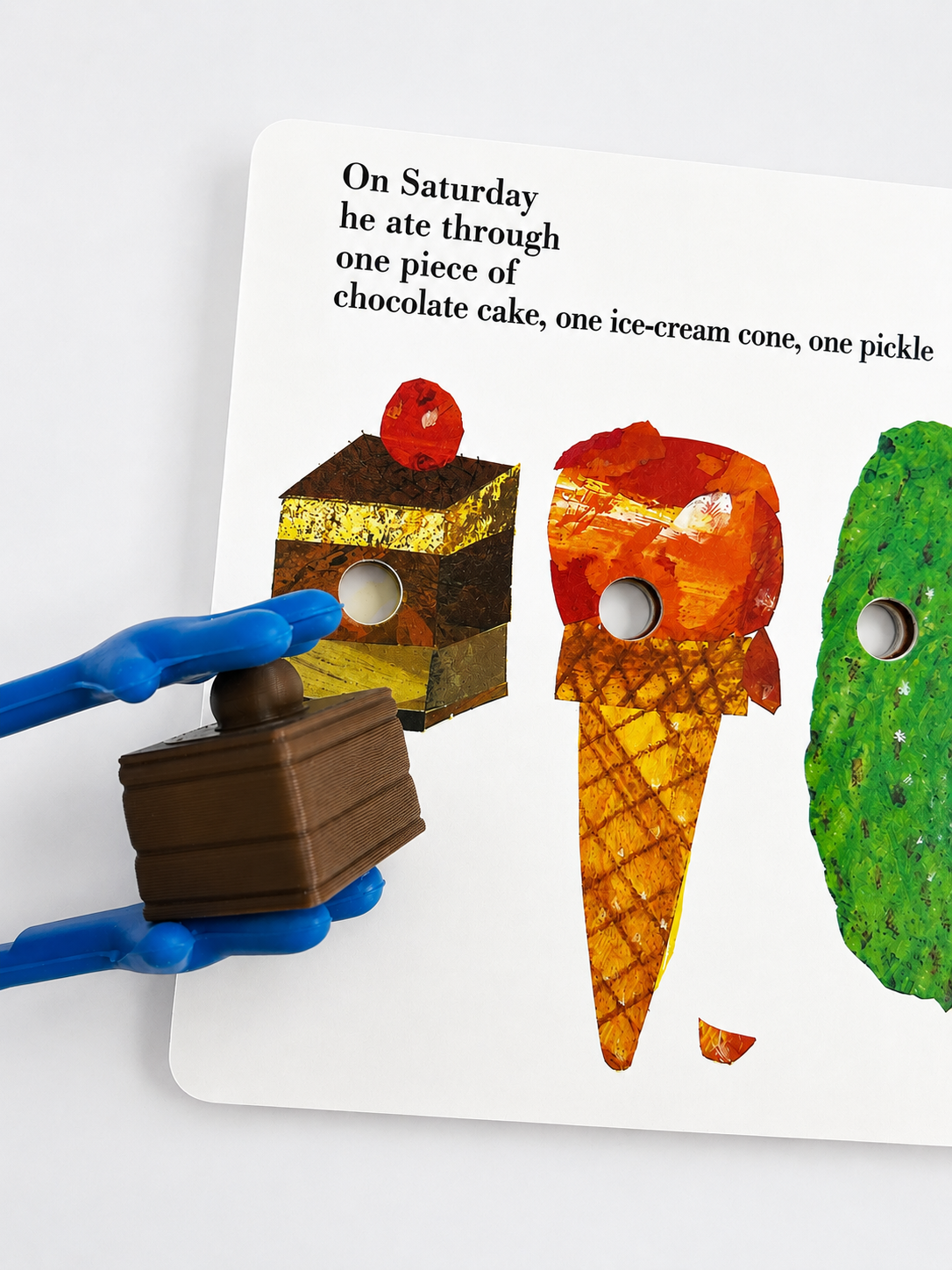 A children's interactive book page showing a story about eating dessert, with cutout illustrations of chocolate cake, an ice cream cone, and a pickle. A blue-handled toy piece is being held in front of the page on a white background.