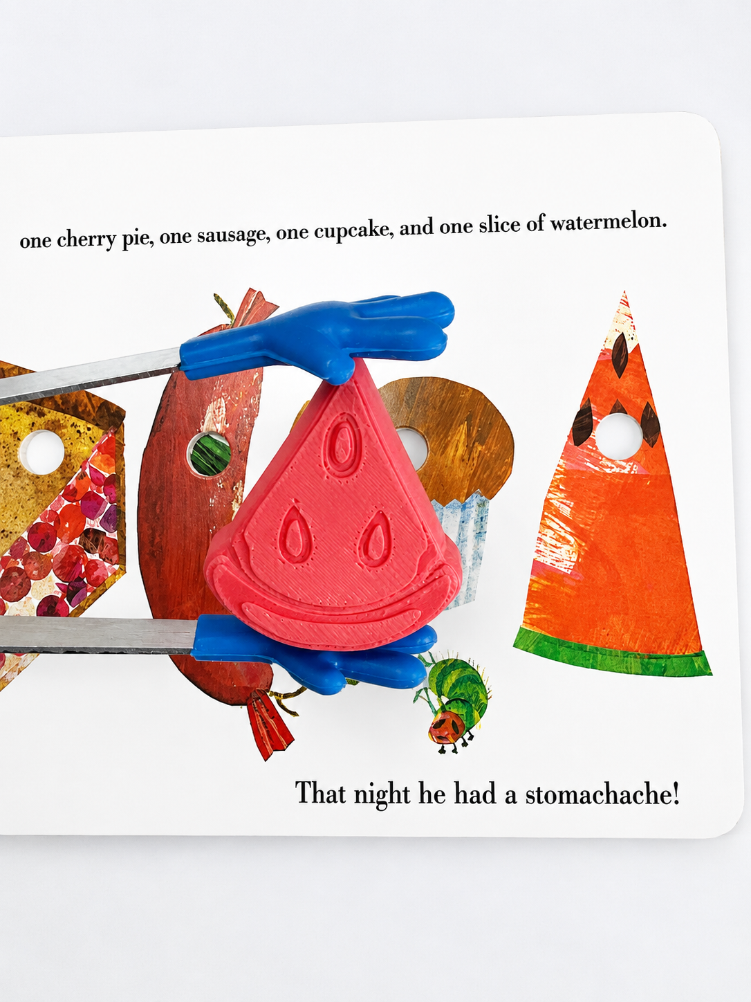 A children's book or activity card showing a watermelon slice puzzle piece being held in blue-handled tweezers. The white card includes printed text and other illustrated food pieces, including a watermelon slice, cherry pie, sausage, and cupcake.