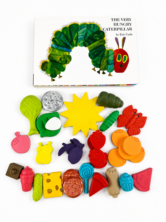 The Very Hungry Caterpillar children's activity book and matching craft set arranged on a white background. The book shows the caterpillar illustration, and the colorful clay pieces below include leaves, fruit, a croissant, a butterfly, and other shapes inspired by the book.