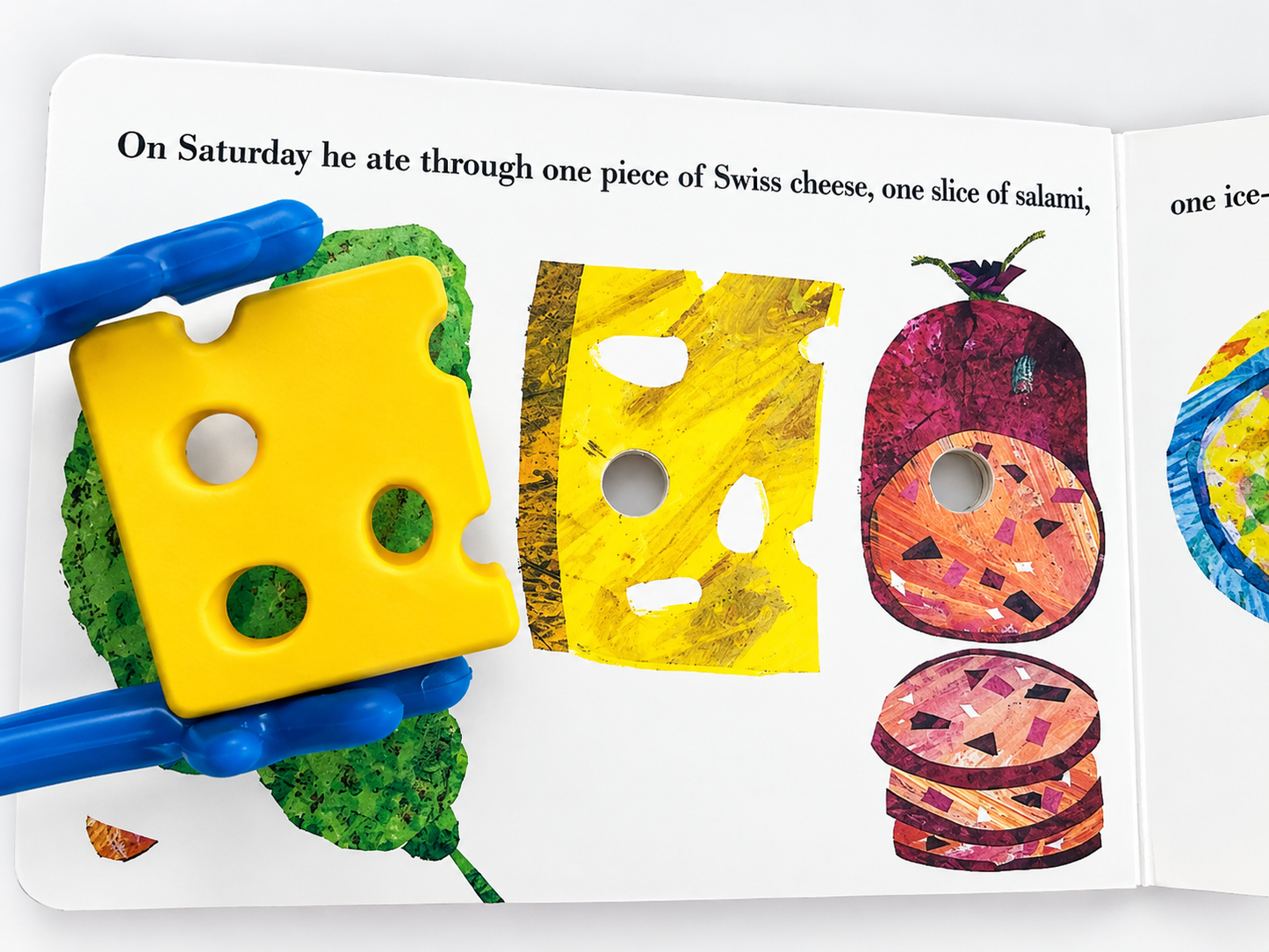 A children's activity book or puzzle page showing illustrated food items and text, with a blue-handled tool holding a yellow cheese-shaped piece over a green textured surface. The page includes drawings of Swiss cheese, a slice of salami, and sliced salami on a white background.