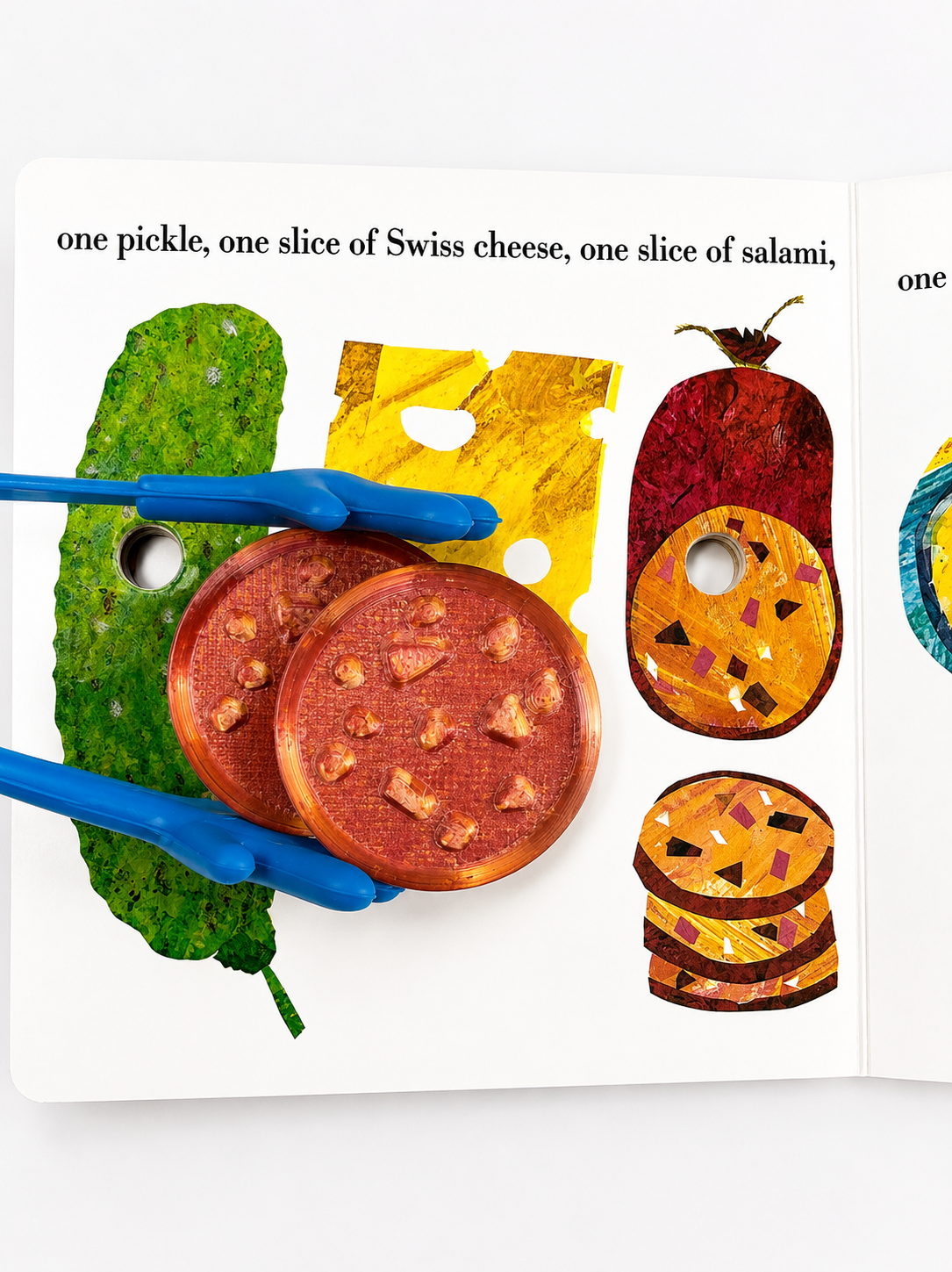 A children's interactive book or activity page showing illustrated food items and blue plastic tongs placed over the pictures on a white page.