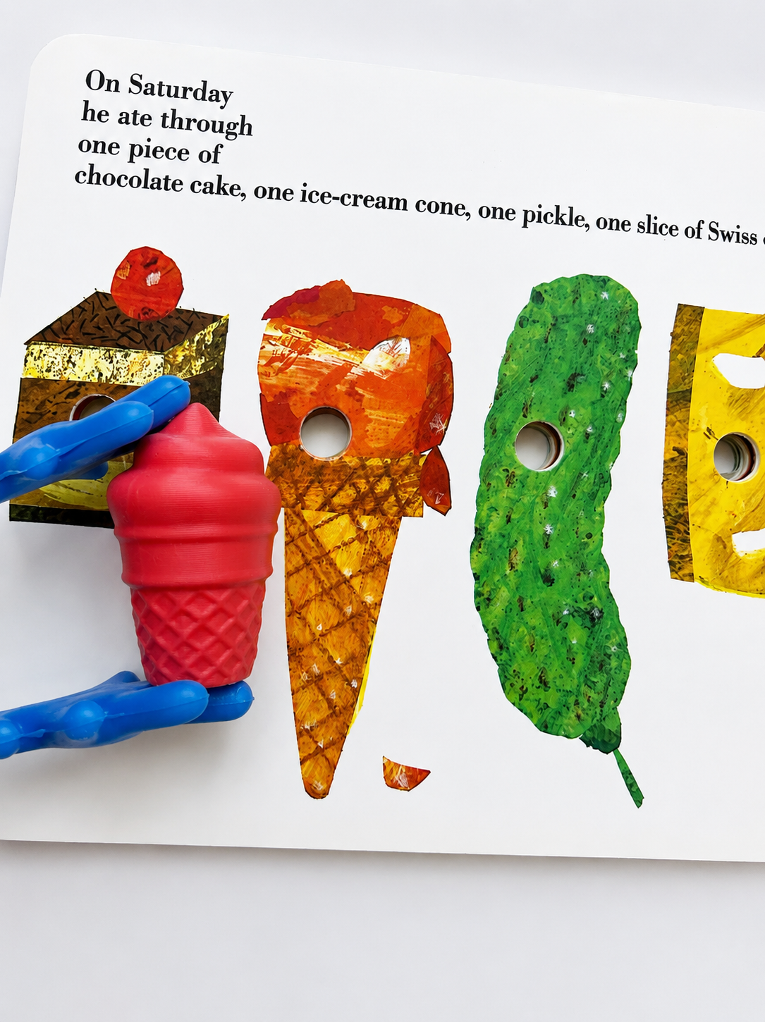 A children's activity card showing a puzzle about what was eaten on Saturday, with illustrated pieces including a chocolate cake, ice cream cone, pickle, and yellow slice. A blue-handled piece is being placed over a red ice cream cone illustration on a white card.