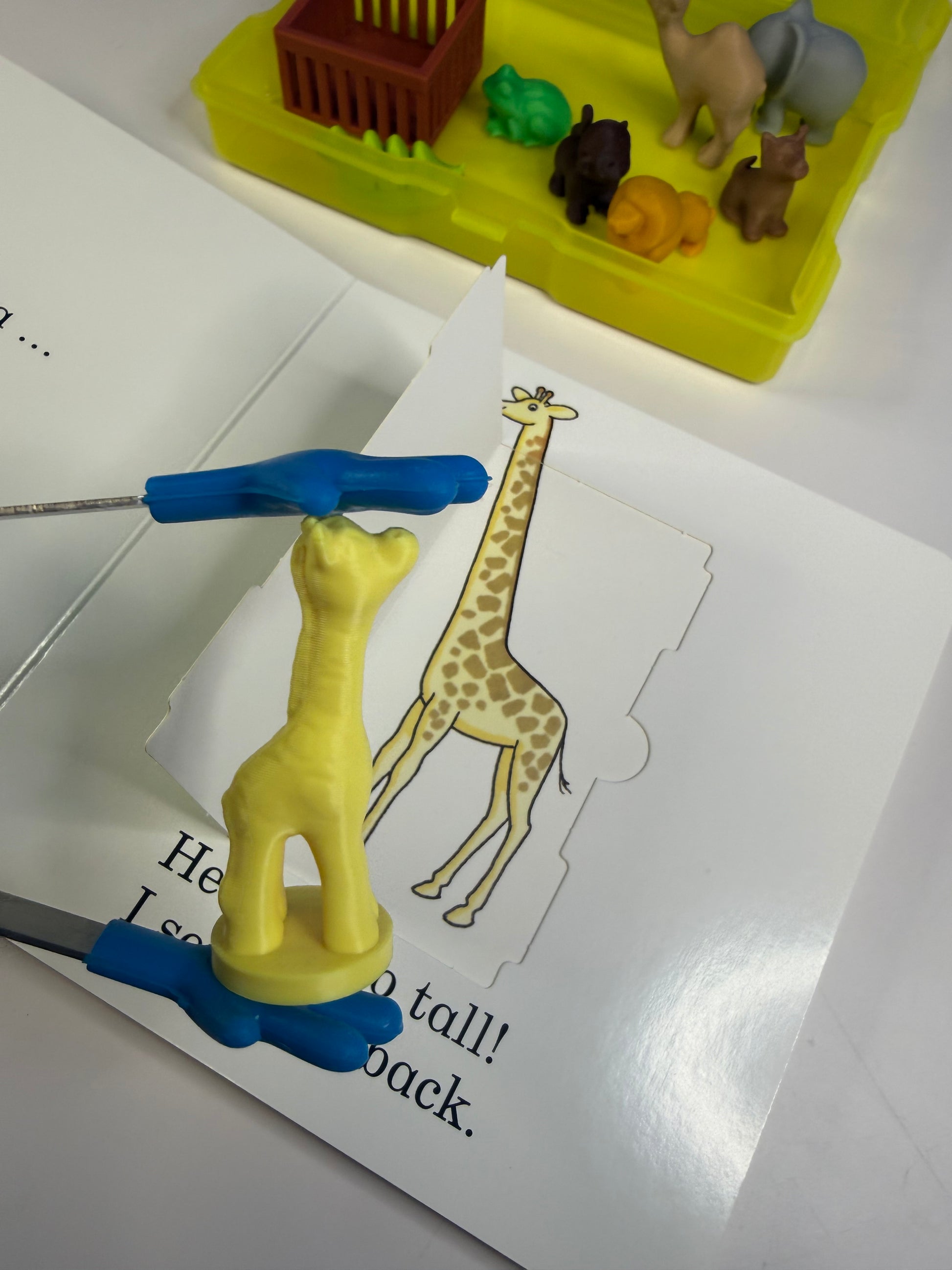 Toy giraffe with a blue base on a book page, surrounded by toy animals in the background.