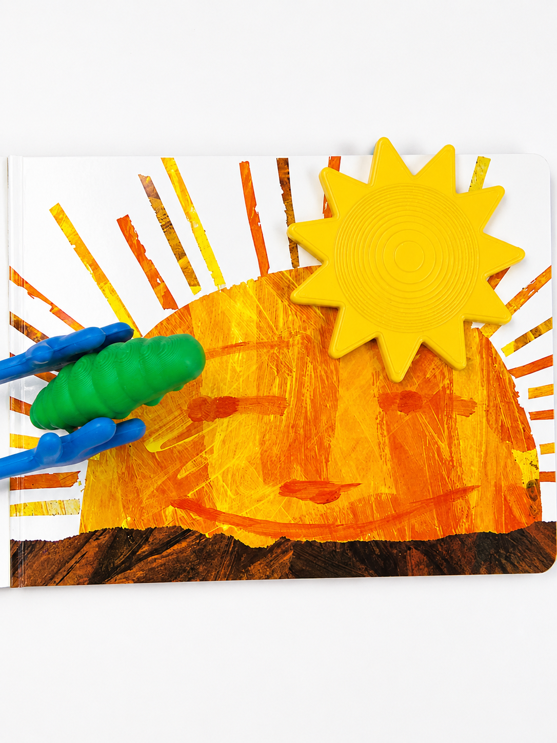 A children's art activity board featuring a child's textured painting of a smiling sun over a brown landscape, with a yellow plastic sun cutout and a blue-and-green plastic sun accessory.